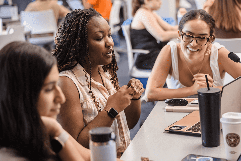 Three female students sit at a table and look at a laptop as they study.